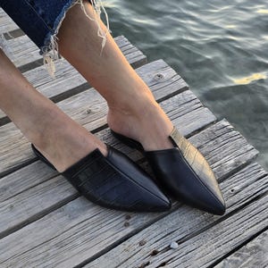 May include: Black leather slip-on shoes with pointed toes. One shoe has a textured, crocodile-like pattern. The shoes are on a wooden dock near the water. The shoes are a size 8.