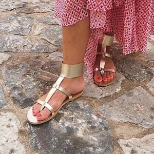 May include: Gold leather sandals with ankle straps and a buckle closure. The sandals have a flat sole and are worn on a woman's feet.
