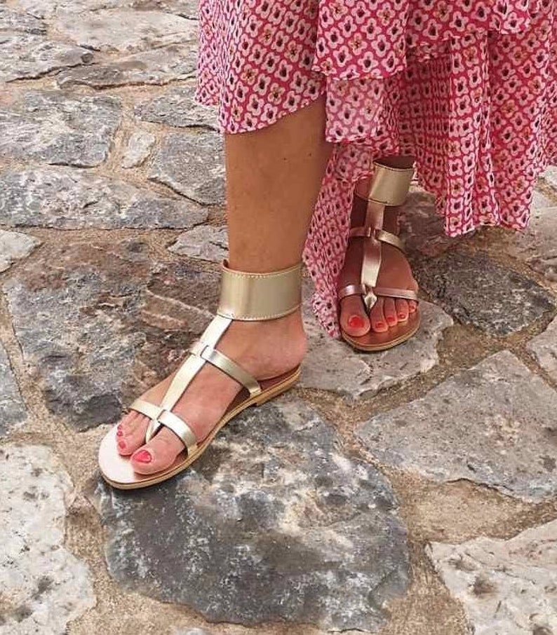 May include: Gold leather sandals with ankle straps and a buckle closure. The sandals have a flat sole and are worn on a woman's feet.