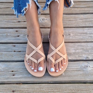 Op de afbeelding: Een paar lichtbruine leren sandalen met gekruiste banden. De sandalen hebben een platte zool en worden gedragen door iemand met witte nagellak.