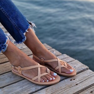 Op de afbeelding: Een paar lichtbruine leren sandalen met gekruiste banden. De sandalen liggen op een houten oppervlak.