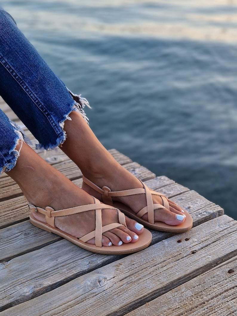 Op de afbeelding: Een paar lichtbruine leren sandalen met gekruiste banden. De sandalen liggen op een houten oppervlak.