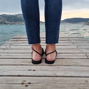 May include: A pair of black leather sandals with thin straps. The sandals are worn by a person standing on a wooden dock.