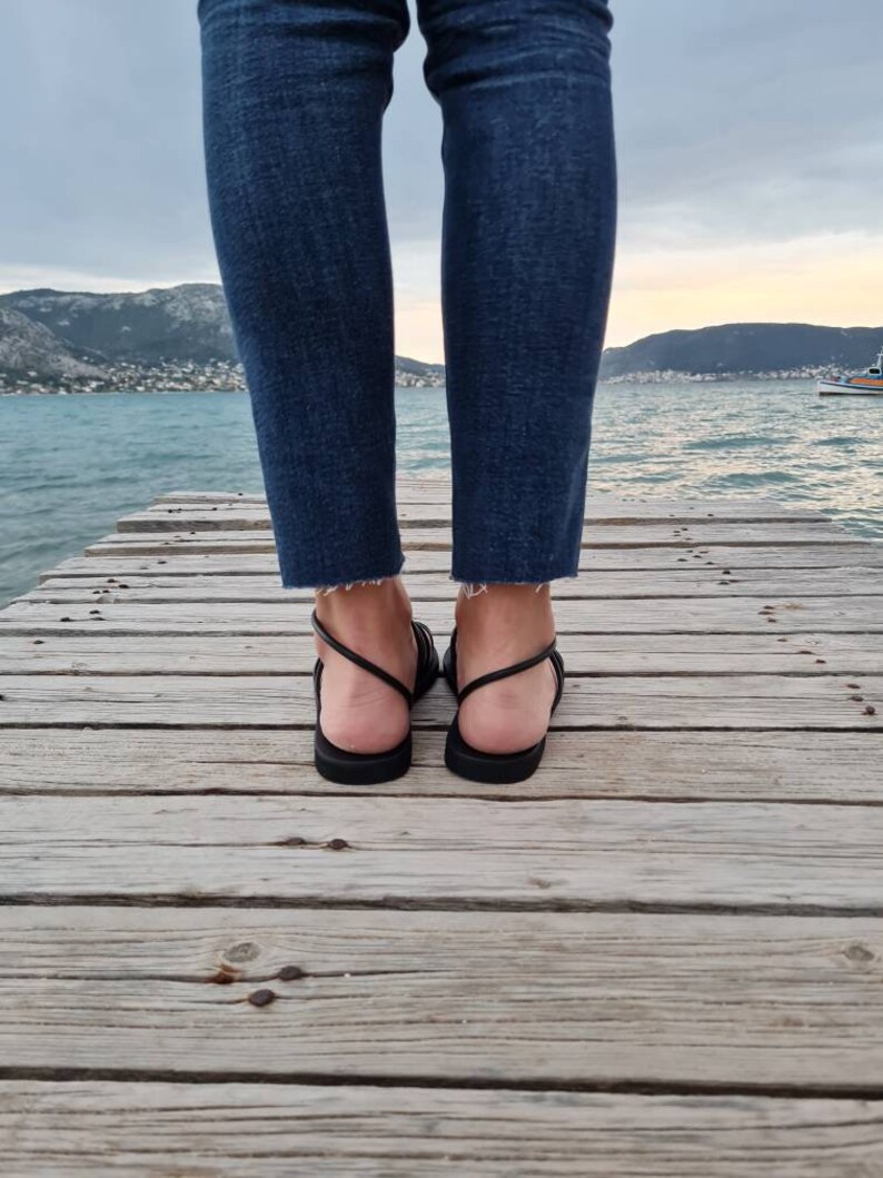 May include: A pair of black leather sandals with thin straps. The sandals are worn by a person standing on a wooden dock.