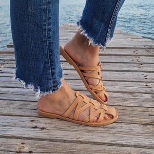 May include: Tan leather sandals with a woven strap design. The sandals have a flat sole and are worn on a wooden surface.