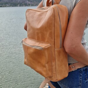 May include: A tan leather backpack with a zippered front pocket and adjustable straps. The backpack is worn by a person standing on a wooden dock.