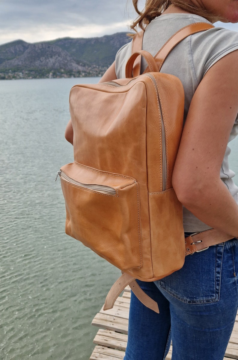 May include: A tan leather backpack with a zippered front pocket and adjustable straps. The backpack is worn by a person standing on a wooden dock.