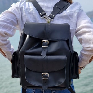 Puede incluir: Una mochila de cuero negro con dos bolsillos grandes en la parte delantera y correas ajustables. La mochila tiene dos hebillas en la parte delantera y una hebilla plateada en la parte superior.