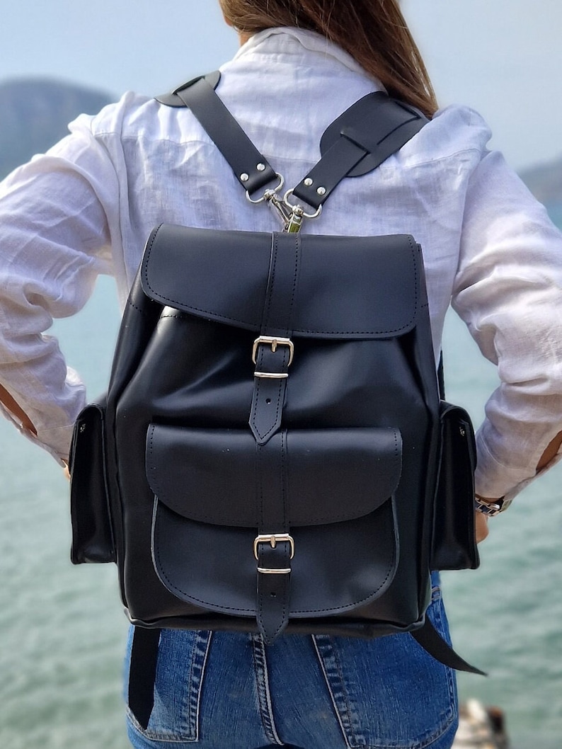 Puede incluir: Una mochila de cuero negro con dos bolsillos grandes en la parte delantera y correas ajustables. La mochila tiene dos hebillas en la parte delantera y una hebilla plateada en la parte superior.