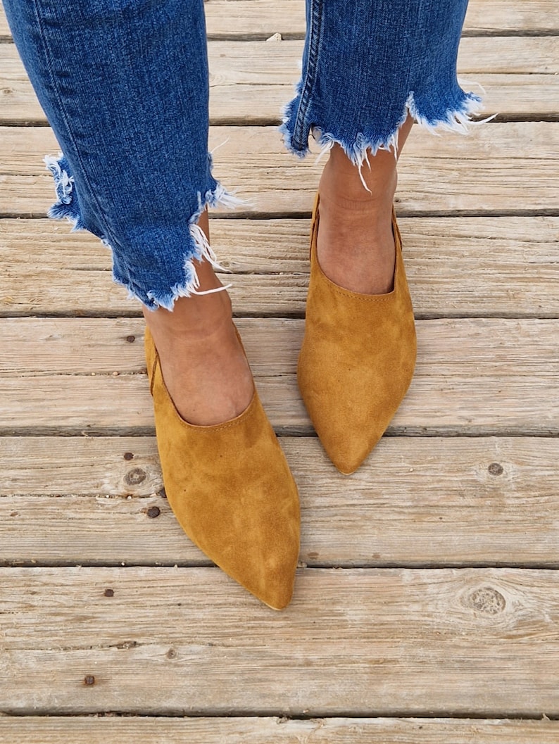 May include: A pair of brown suede pointed-toe flats with a low heel. The shoes are worn by a person wearing blue jeans.