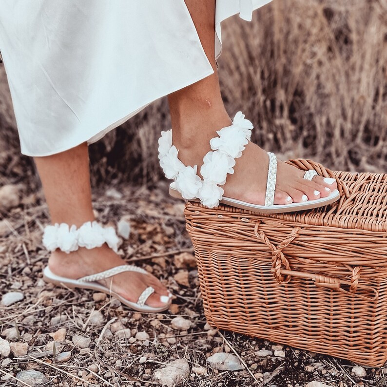 May include: White sandals with a floral design and a beaded strap. The sandals are on a wicker basket.