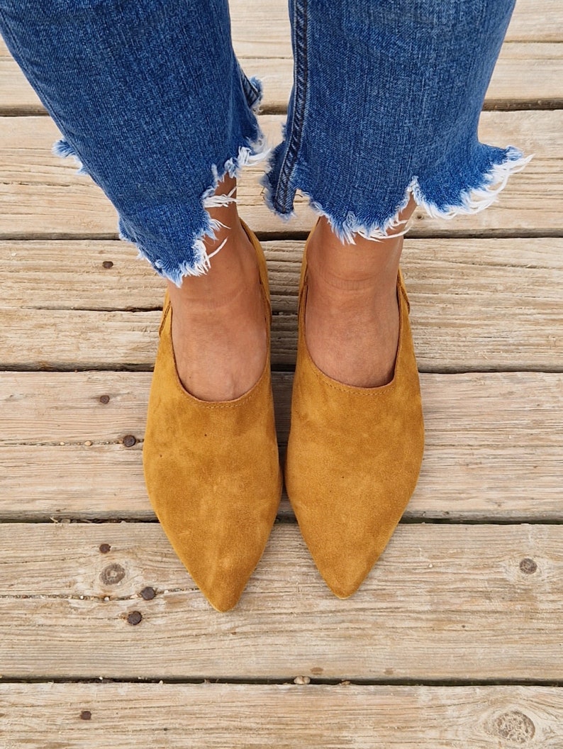 May include: A pair of brown suede pointed-toe flats with a low heel. The shoes are worn by a person wearing blue jeans with frayed hems.