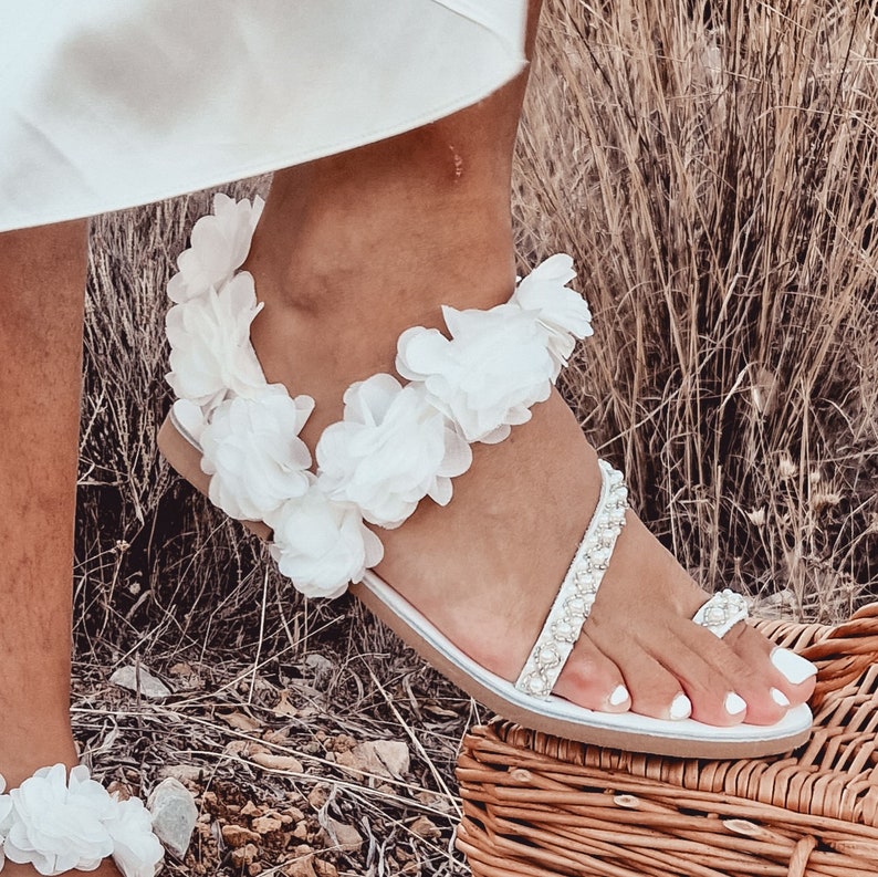 May include: White sandals with a flower embellishment around the ankle strap. The sandals have a beaded strap across the foot and a toe ring.