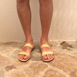 May include: A pair of light brown leather sandals with two straps and a buckle closure. The sandals are worn on a person's feet, standing on a stone floor.