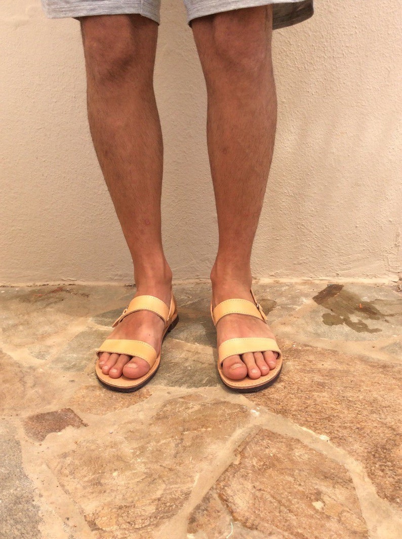 May include: A pair of light brown leather sandals with two straps and a buckle closure. The sandals are worn on a person's feet, standing on a stone floor.