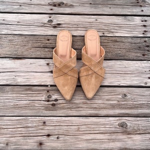 May include: A pair of light brown suede heeled sandals with criss-cross straps. The sandals are on a wooden surface.