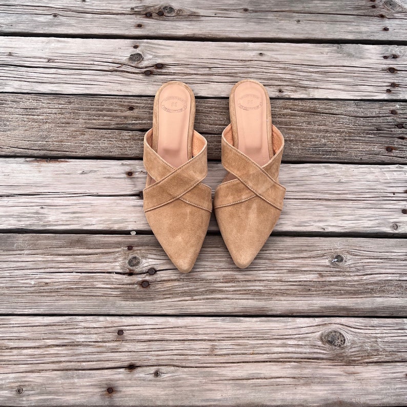 May include: A pair of light brown suede heeled sandals with criss-cross straps. The sandals are on a wooden surface.
