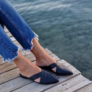 May include: A pair of black leather mules with a criss-cross strap design. The shoes have a pointed toe and are worn by a person sitting on a wooden dock.