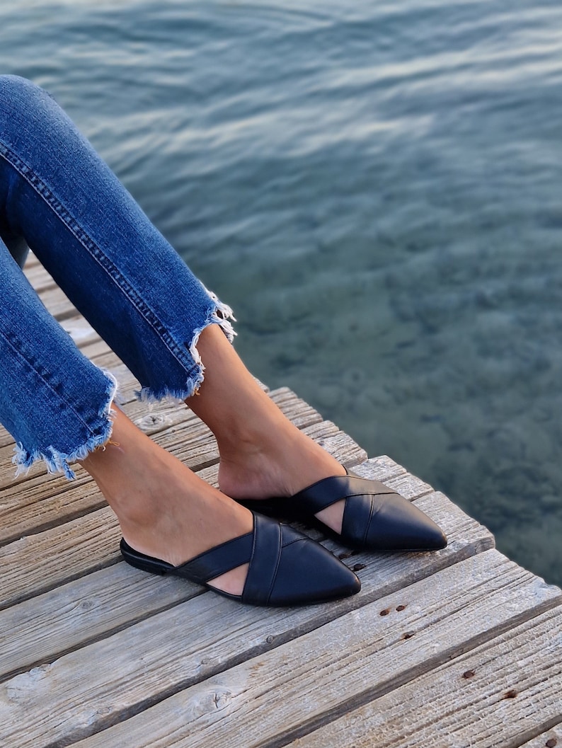 May include: A pair of black leather mules with a criss-cross strap design. The shoes have a pointed toe and are worn by a person sitting on a wooden dock.