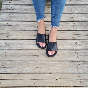 May include: A pair of black leather slide sandals with a simple design. The sandals are worn by a person with bare feet and are on a wooden surface.