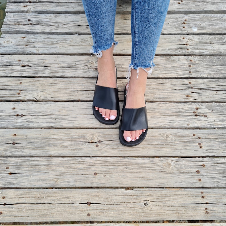 May include: A pair of black leather slide sandals with a simple design. The sandals are worn by a person with bare feet and are on a wooden surface.