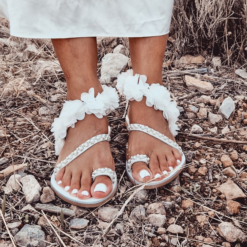 May include: White sandals with a flower embellishment on the ankle strap. The sandals have a beaded design and are worn on a person's feet.