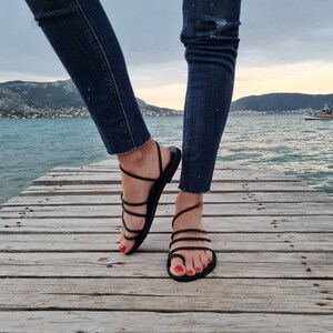 May include: A pair of black leather sandals with multiple straps wrapping around the ankles and feet. The sandals are worn by a person standing on a wooden dock.