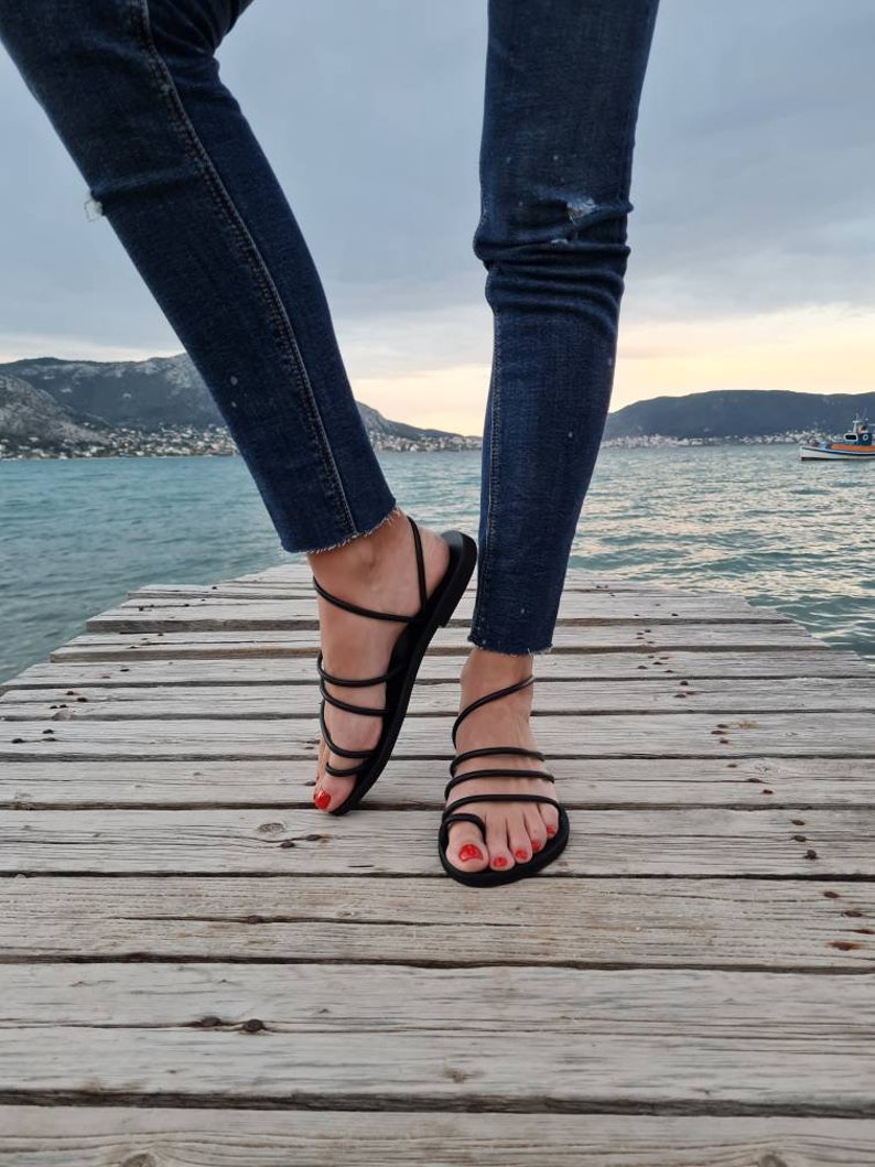 May include: A pair of black leather sandals with multiple straps wrapping around the ankles and feet. The sandals are worn by a person standing on a wooden dock.
