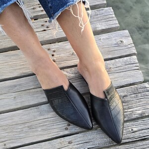 May include: Black leather slip-on mules with a pointed toe and textured vamp. The shoes are on a wooden dock, with the water visible in the background. The mules have a crocodile-like embossed pattern.