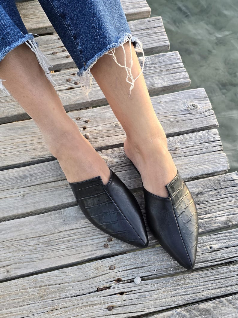 May include: Black leather slip-on mules with a pointed toe and textured vamp. The shoes are on a wooden dock, with the water visible in the background. The mules have a crocodile-like embossed pattern.