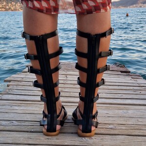 Op de afbeelding: Zwarte gladiatorsandalen van leer met meerdere banden en gespen. De sandalen hebben een rits aan de achterkant en worden gedragen op een houten pier.