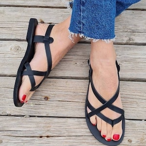 Op de afbeelding: Zwarte leren sandalen met gekruiste banden en een teenriem. De sandalen worden gedragen op een houten oppervlak.