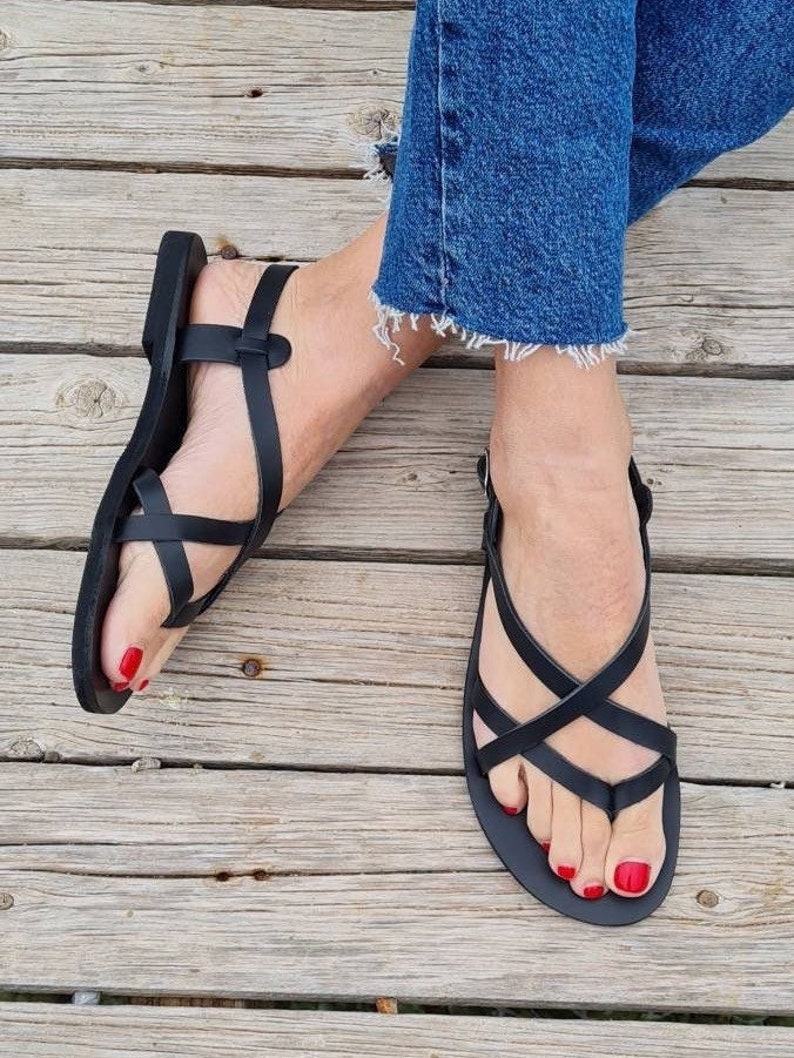Op de afbeelding: Zwarte leren sandalen met gekruiste banden en een teenriem. De sandalen worden gedragen op een houten oppervlak.