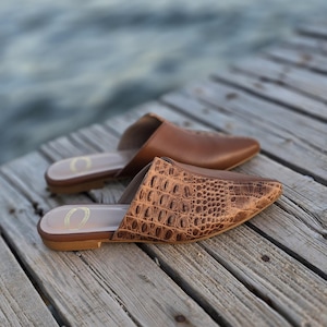 May include: Two pairs of brown leather mules are displayed on a wooden surface. One pair features a textured, crocodile-like pattern, while the other has a smooth finish. The shoes have a pointed toe design and are open-backed.