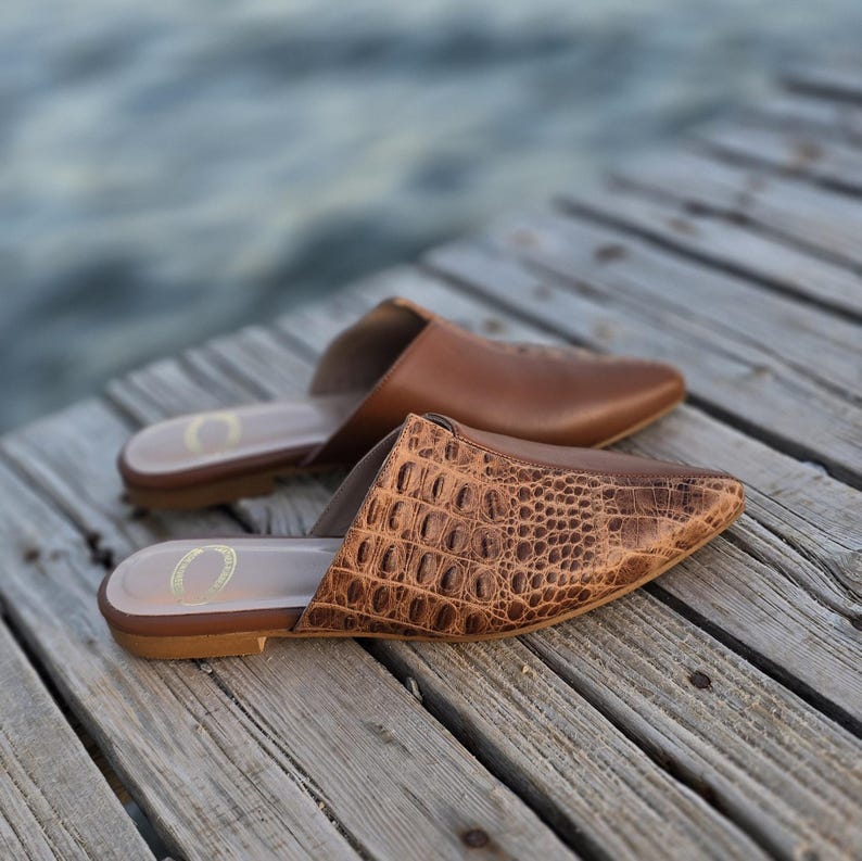 May include: Two pairs of brown leather mules are displayed on a wooden surface. One pair features a textured, crocodile-like pattern, while the other has a smooth finish. The shoes have a pointed toe design and are open-backed.