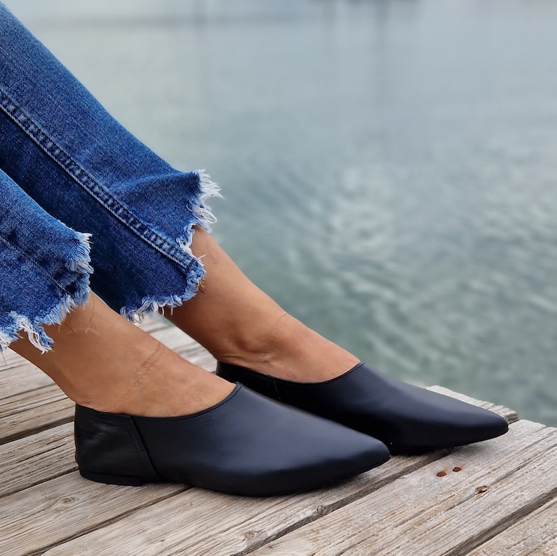 May include: A pair of black leather flats with pointed toes. The shoes are worn by a person sitting on a wooden dock.