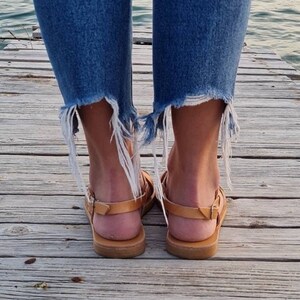 Op de afbeelding: Een paar bruine leren sandalen met een gekruist bandontwerp. De sandalen worden gedragen door iemand die op een houten pier staat.