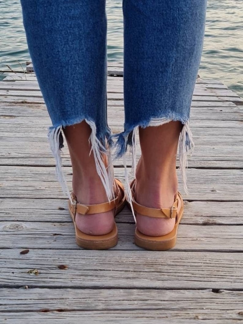 Op de afbeelding: Een paar bruine leren sandalen met een gekruist bandontwerp. De sandalen worden gedragen door iemand die op een houten pier staat.