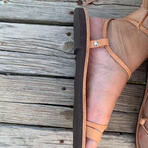 May include: Tan leather sandals with a dark brown sole. The sandals have multiple straps that wrap around the foot and ankle. The sandals are shown on a weathered wooden surface.