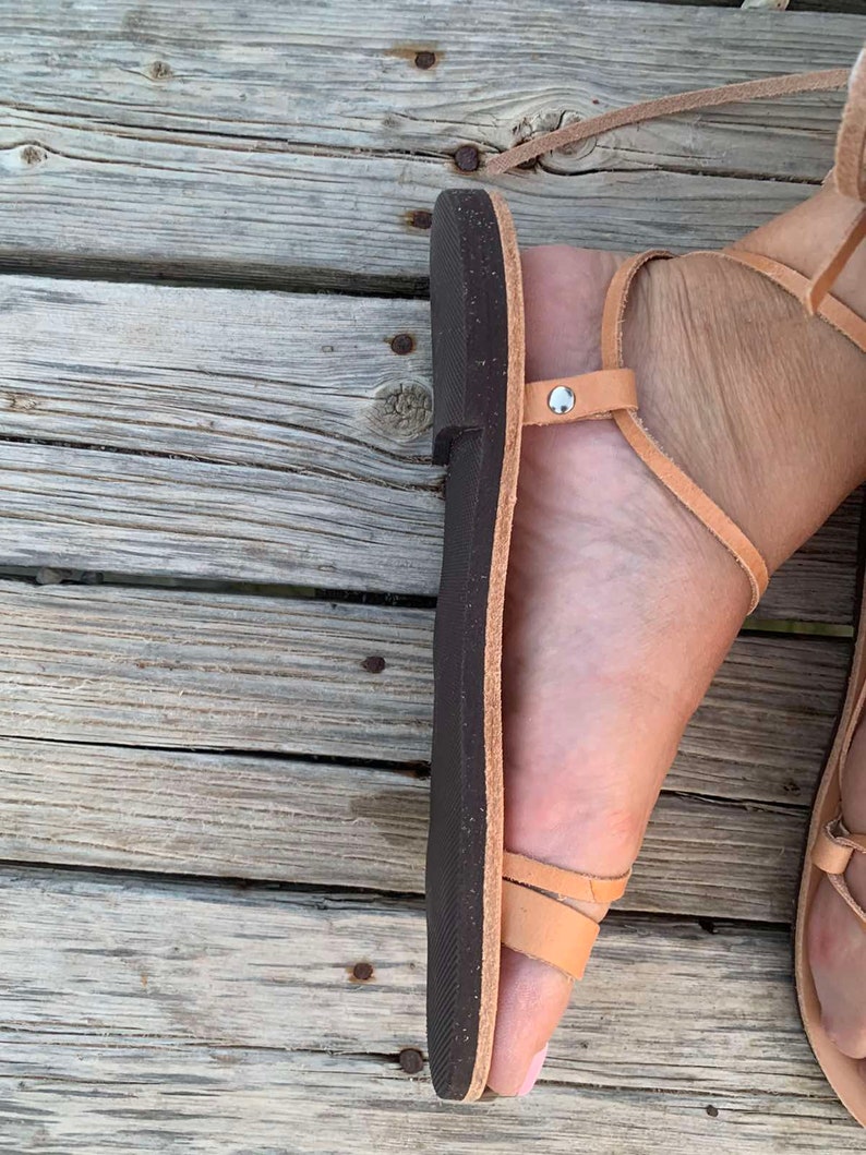 May include: Tan leather sandals with a dark brown sole. The sandals have multiple straps that wrap around the foot and ankle. The sandals are shown on a weathered wooden surface.