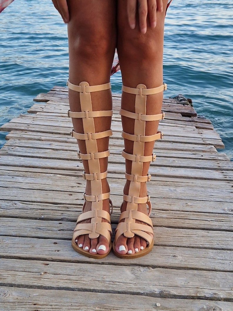 Op de afbeelding: Een paar bruine leren gladiatorsandalen met meerdere banden en gespen. De sandalen worden gedragen op een houten pier.
