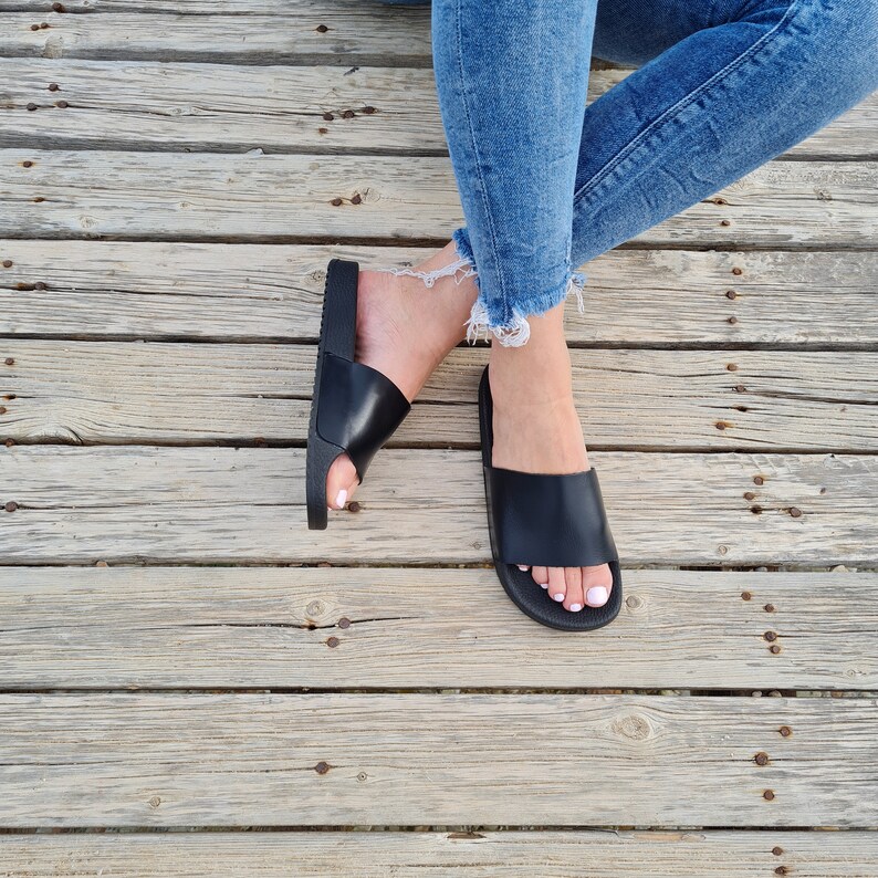 May include: A pair of black leather slide sandals with a simple design. The sandals are worn by a person with denim jeans.