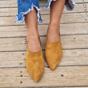 May include: A pair of brown suede pointed-toe flats. The shoes are worn by a person wearing blue jeans.