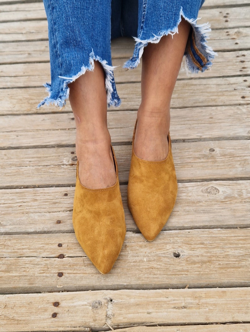 May include: A pair of brown suede pointed-toe flats. The shoes are worn by a person wearing blue jeans.