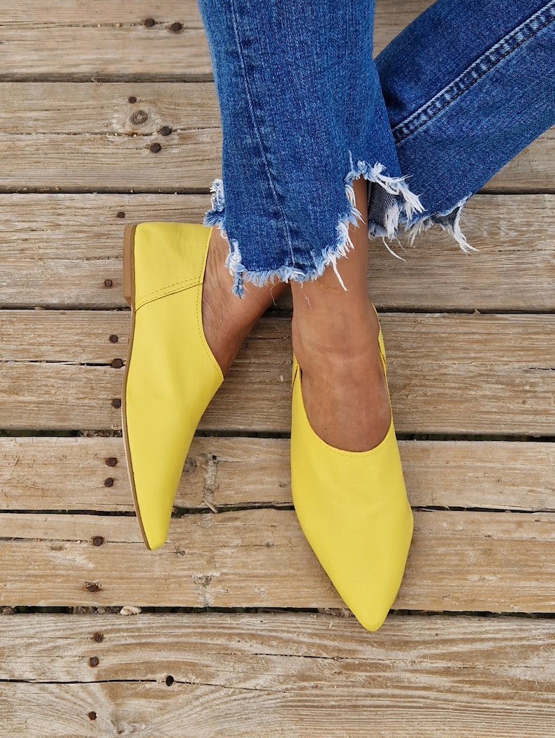 May include: A pair of bright yellow leather flats with pointed toes. The shoes are worn by a person wearing blue jeans.