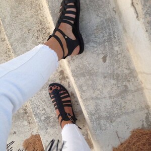 May include: A pair of black leather gladiator sandals with straps that wrap around the ankles. The sandals are worn on a person's feet, with the person wearing white jeans.