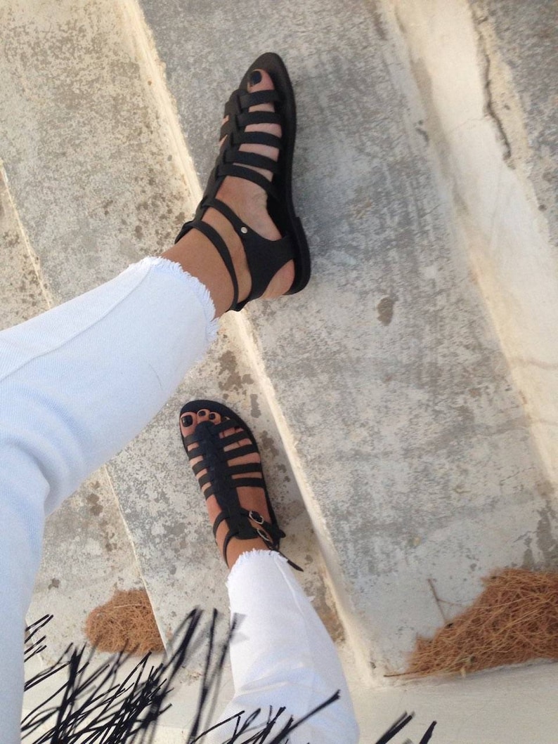 May include: A pair of black leather gladiator sandals with straps that wrap around the ankles. The sandals are worn on a person's feet, with the person wearing white jeans.