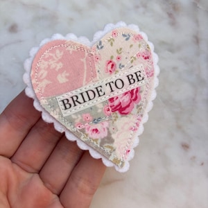 Puede incluir: Un parche de tela en forma de corazón con un borde festoneado blanco. El parche está hecho de tela rosa y con estampado floral. Las palabras "BRIDE TO BE" están impresas en una pancarta blanca en el centro del corazón.