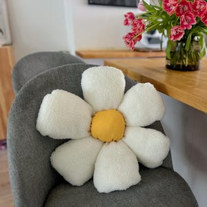 May include: A white plush daisy-shaped cushion with a yellow centre, sitting on a grey chair. The cushion is a home decor item.