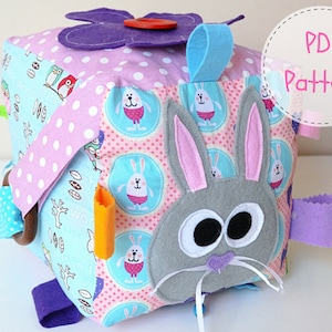 May include: A colourful fabric cube toy with a bunny face, a flower, and a variety of textures. The cube has a pink and white polka dot fabric, a blue fabric with white owls, and a pink fabric with white bunnies. The toy is designed for babies and toddlers.